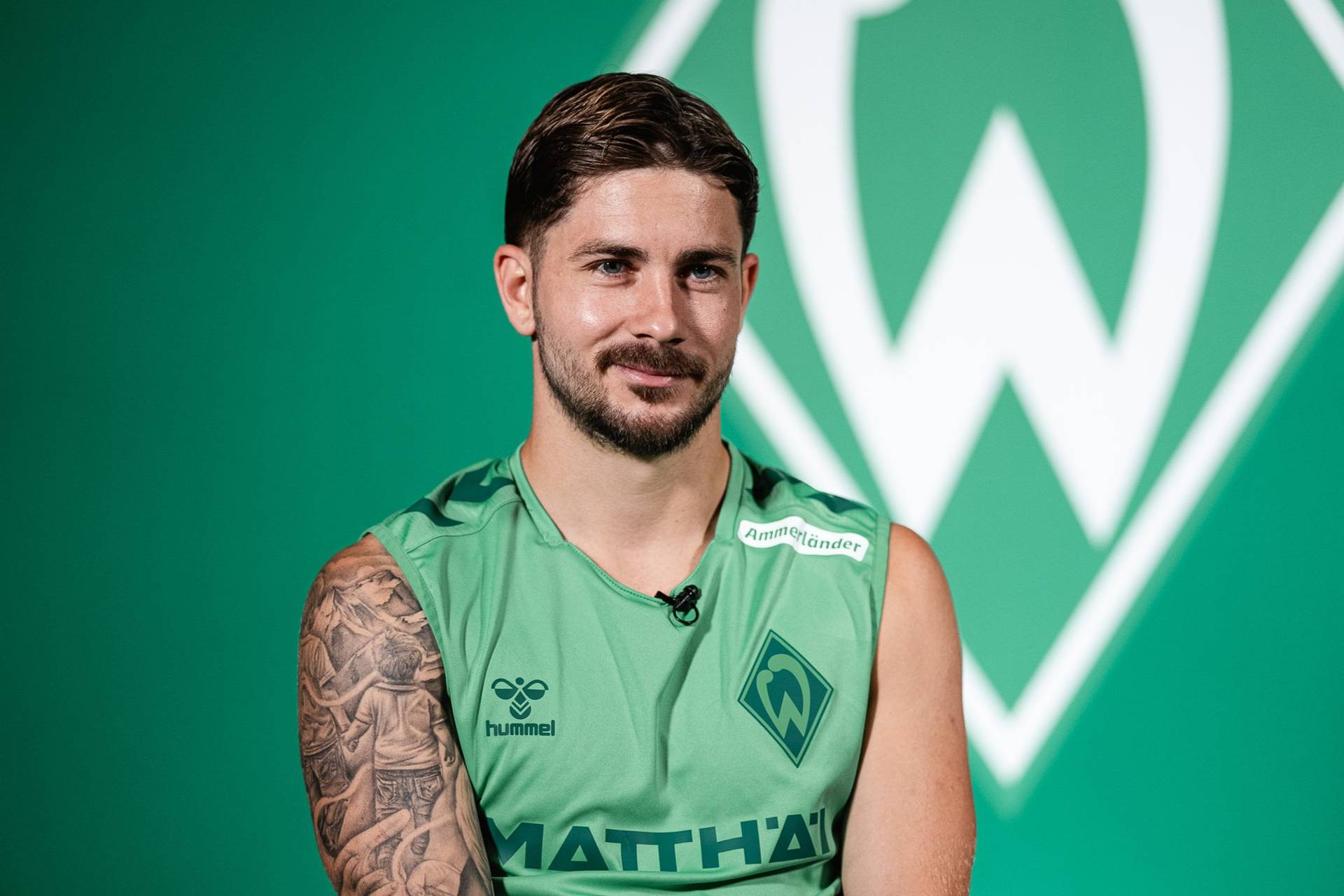 Eine Person in einem grünen Sporttrikot mit einem tätowierten Arm sitzt vor einem grünen Hintergrund, auf dem ein weiß-grünes Emblem zu sehen ist.