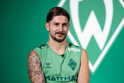 Eine Person in einem grünen Sporttrikot mit einem tätowierten Arm sitzt vor einem grünen Hintergrund, auf dem ein weiß-grünes Emblem zu sehen ist.