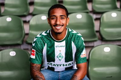 Ein lächelnder Mann in einem grün-weiß gestreiften Fußballtrikot sitzt auf einem Stadionplatz, umgeben von leeren grünen Sitzen.