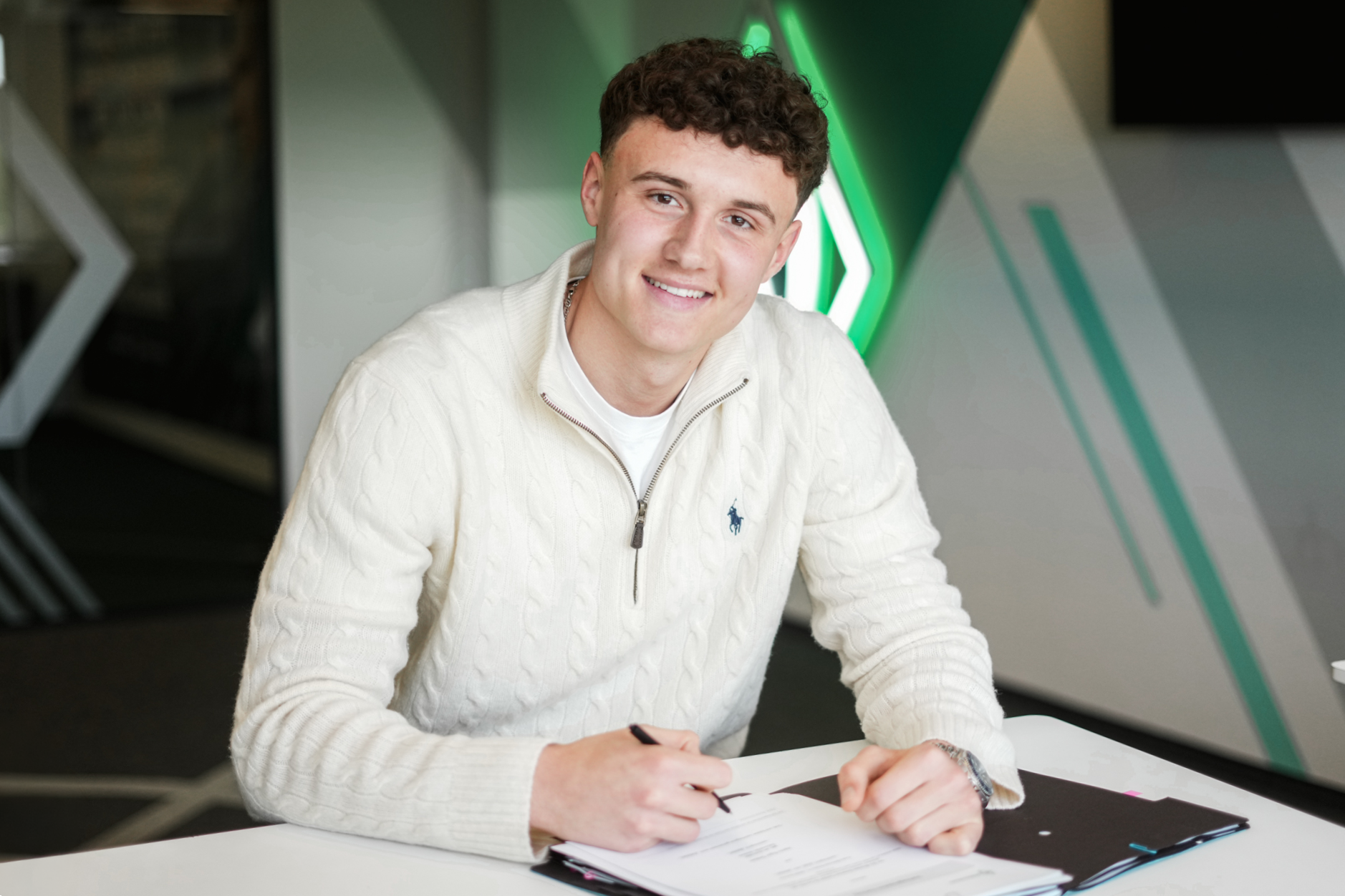 Egor Greber sitzt mit einem Stift in der Hand an einem Tisch, während hinter ihm eine Werder-Raute leuchtet.