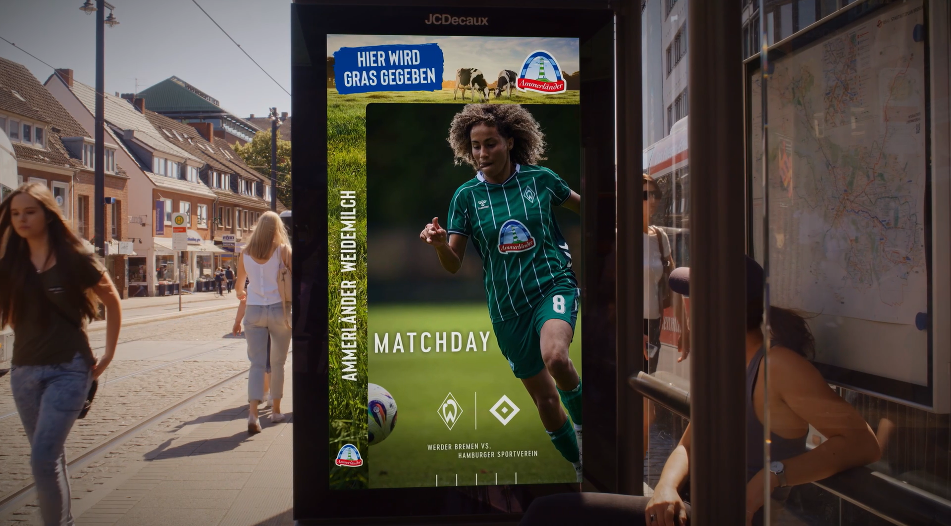 Eine digitale Werbetafel mit einer Fußballspielerin in einem grünen Trikot, im Hintergrund eine städtische Straßenkulisse und Passanten.