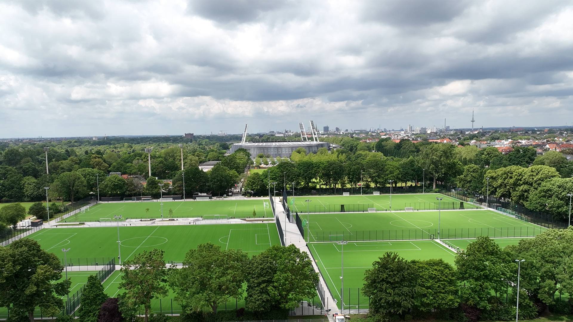 Luftaufnahme mehrerer grüner Fußballplätze, umgeben von Bäumen, mit einer Stadtkulisse und bewölktem Himmel im Hintergrund.