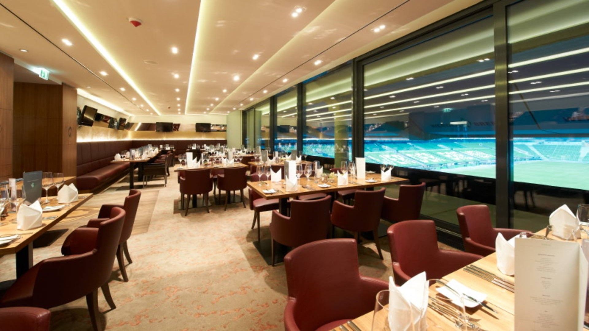 Elegantes Restaurant mit ordentlich gedeckten Tischen, roten Stühlen und großen Fenstern mit Blick auf ein Stadion. Warme Beleuchtung schafft eine gemütliche Atmosphäre.