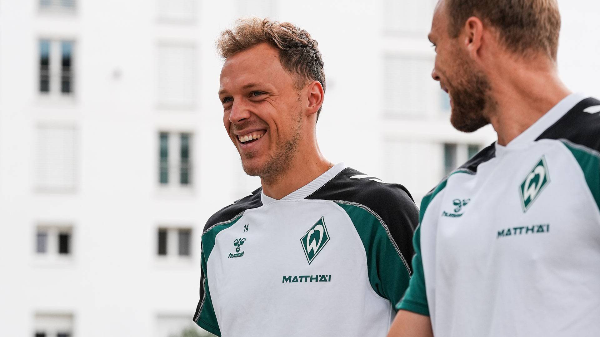 Zwei Fußballspieler in grün-weißen Trikots lächeln, während sie im Freien spazieren gehen; im Hintergrund ist ein Gebäude zu sehen.