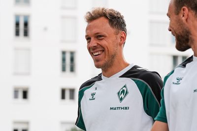 Zwei Fußballspieler in grün-weißen Trikots lächeln, während sie im Freien spazieren gehen; im Hintergrund ist ein Gebäude zu sehen.