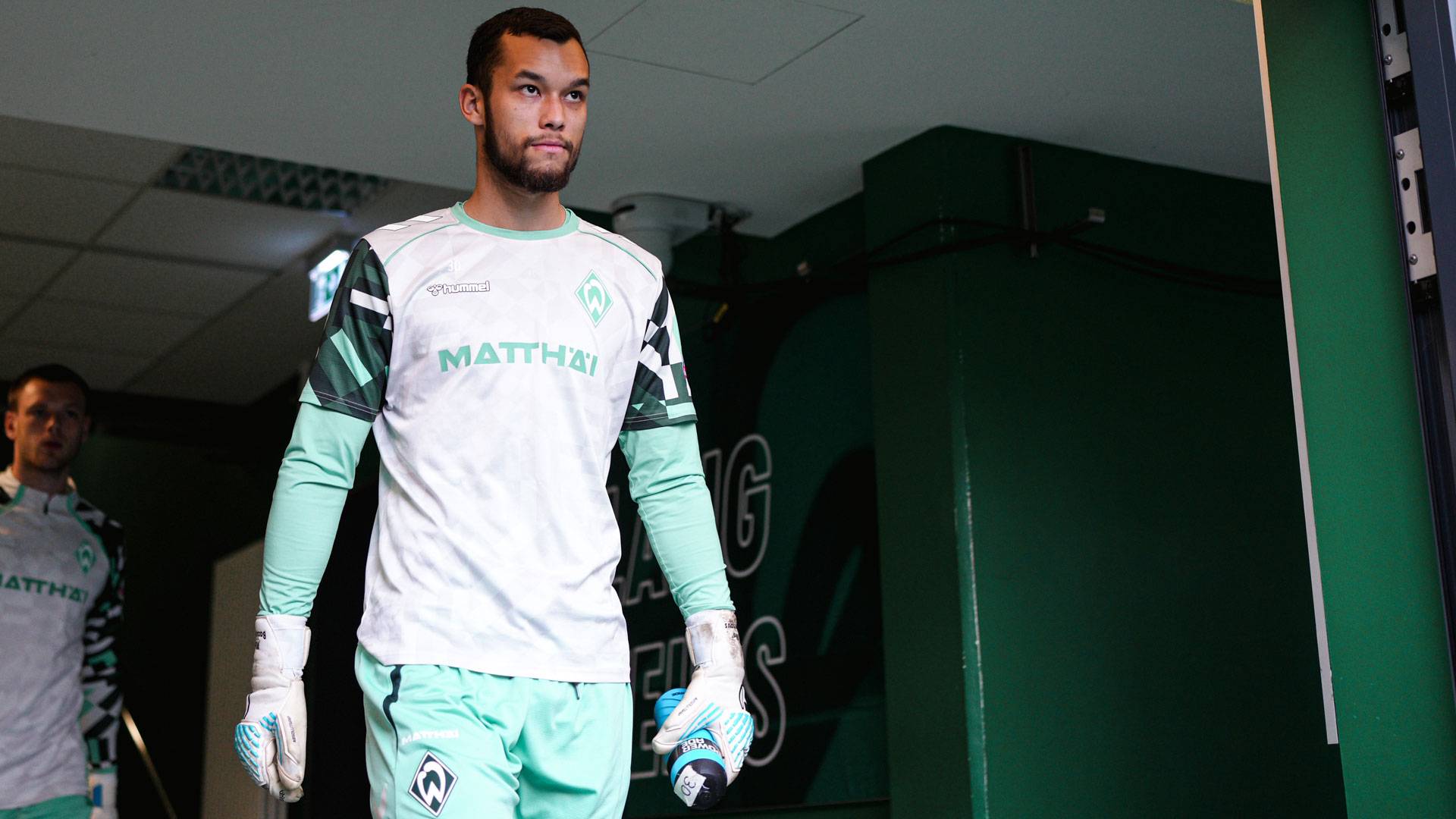 Ein Fußballspieler in einem grün-weißen Trikot geht mit einer Wasserflasche in der Hand einen Flur entlang; hinter ihm ist ein weiterer Spieler zu sehen.