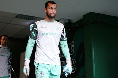 Ein Fußballspieler in einem grün-weißen Trikot geht mit einer Wasserflasche in der Hand einen Flur entlang; hinter ihm ist ein weiterer Spieler zu sehen.