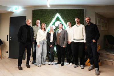 Eine Gruppe von acht Personen steht zusammen in einem Innenraum und lächelt vor einer grünen Wand mit einem beleuchteten dreieckigen Logo. Sie sind leger gekleidet.