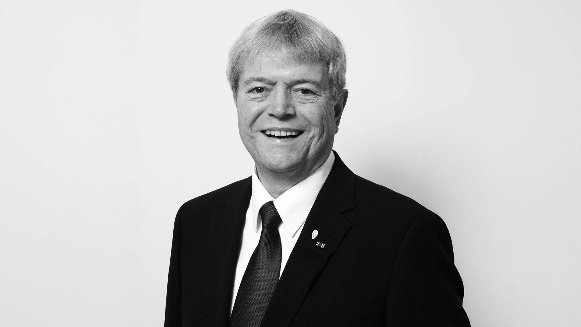 Lächelnder Mann in Anzug und Krawatte, mit einer Anstecknadel am Revers, vor einem schlichten weißen Hintergrund. Schwarz-Weiß-Foto.