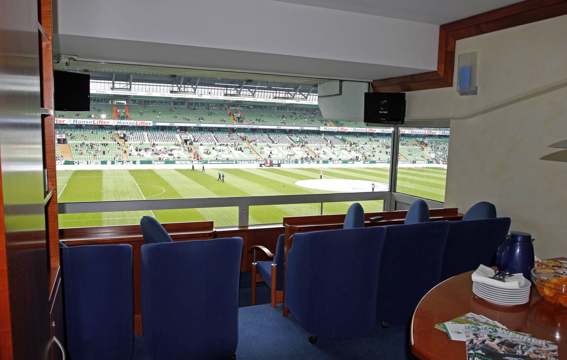 Luxuriöse Stadionloge mit blauen Plüschsitzen und Blick auf ein Fußballfeld. Speisen und Getränke stehen auf einem Tisch im Vordergrund.