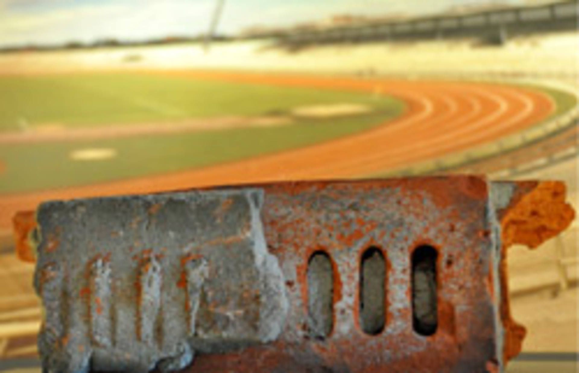 Nahaufnahme eines rostigen Metallträgers mit unscharfem Hintergrund eines Sportstadions mit Laufbahn und leeren Tribünen.