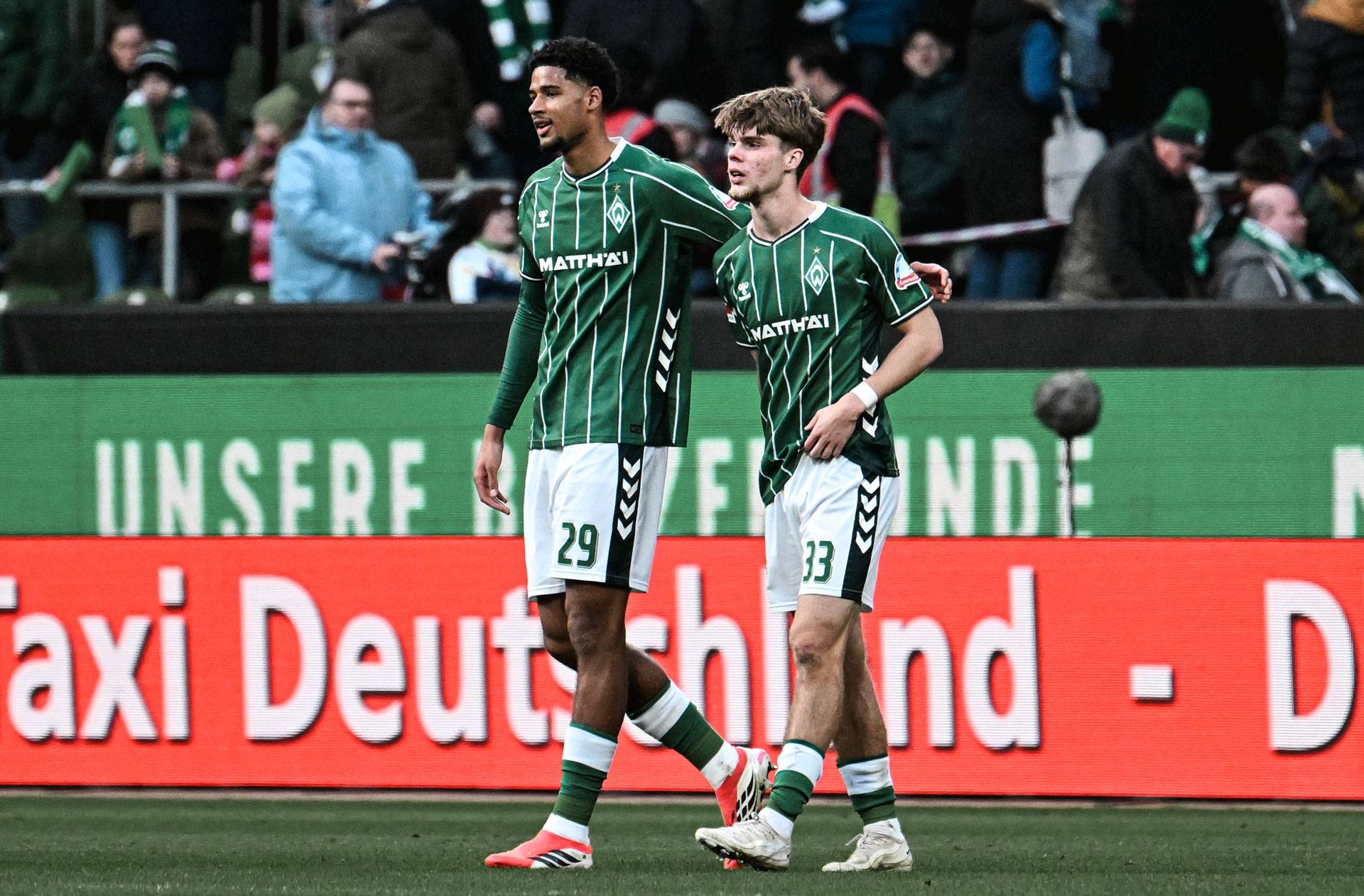 Zwei Fußballspieler in grün-weiß gestreiften Trikots laufen Seite an Seite über den Platz, umgeben von Zuschauern.