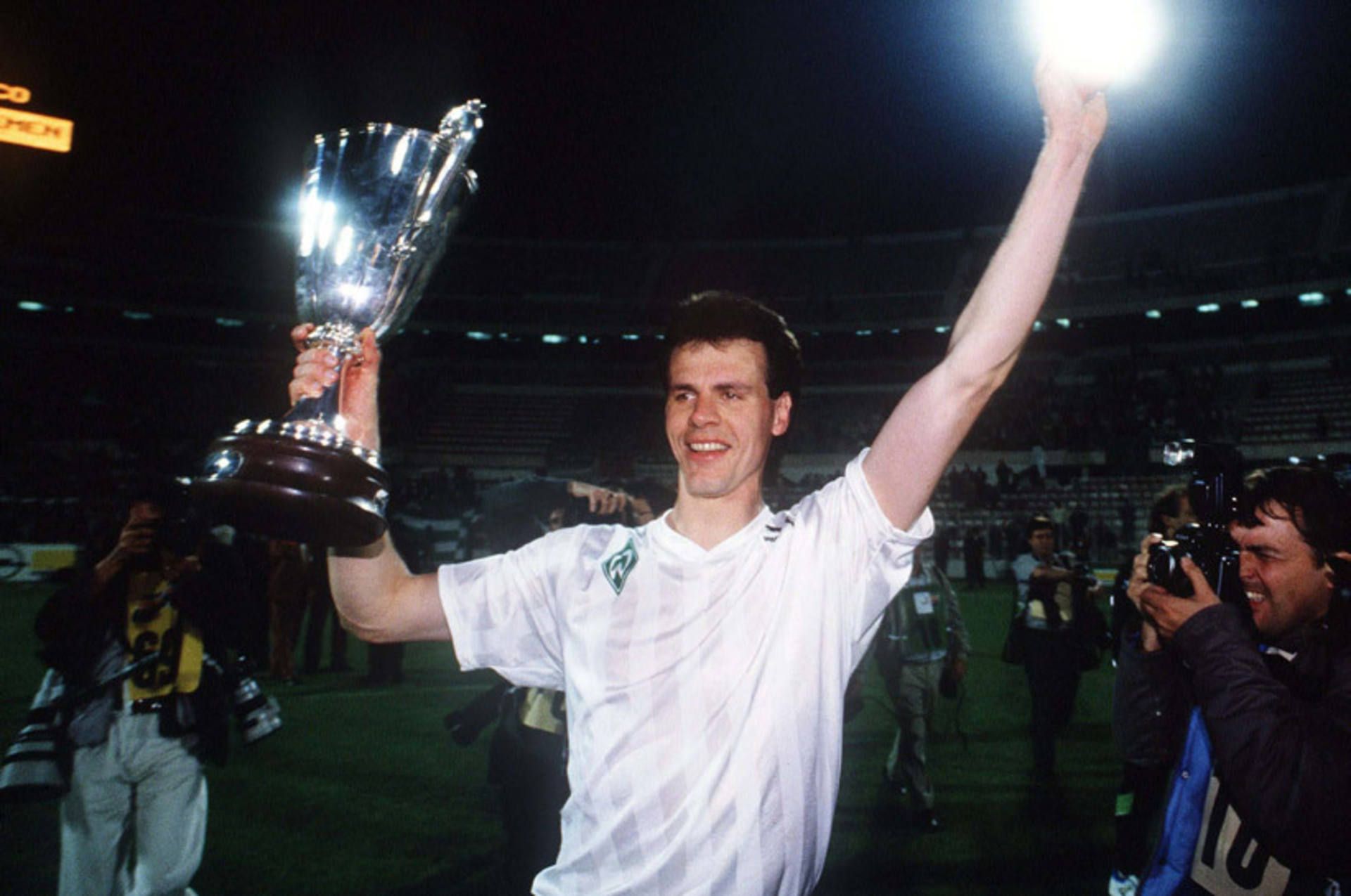 Rune Bratseth in einem weißen Trikot hält triumphierend den Europapokal der Pokalsieger unter den hellen Stadionlichtern in den Händen, umgeben von Fotografen.