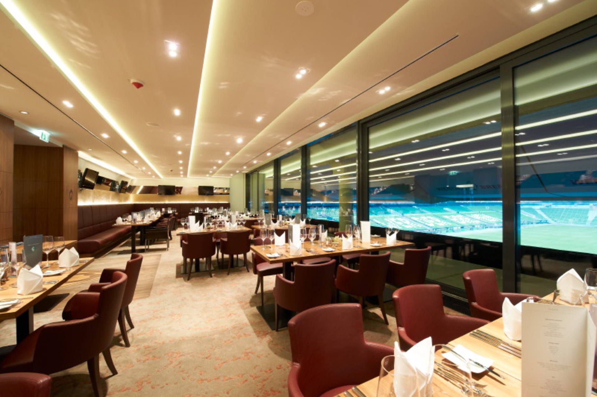 Elegantes Restaurantinterieur mit ordentlich angeordneten Tischen, roten Stühlen und großen Fenstern, die einen Blick auf ein hell erleuchtetes Sportstadion bieten.