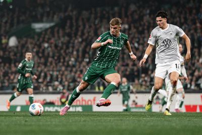 Ein Fußballspieler in grüner Kleidung dribbelt während eines Spiels an einem Gegner in weißer Kleidung vorbei, im Hintergrund ist eine Zuschauermenge zu sehen.