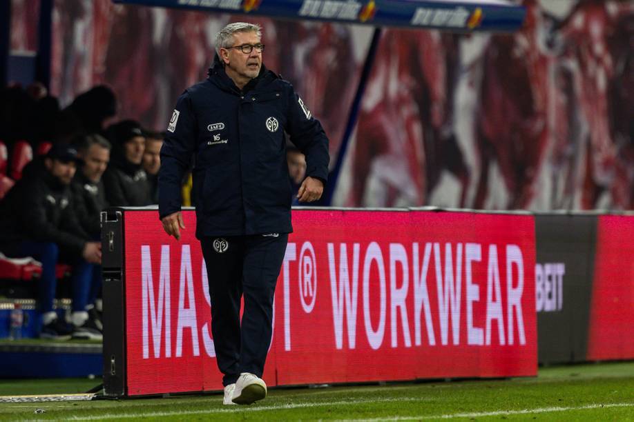 Ein Fußballtrainer in einem marineblauen Trainingsanzug läuft während eines Spiels an der Seitenlinie in der Nähe einer beleuchteten roten Werbetafel entlang.