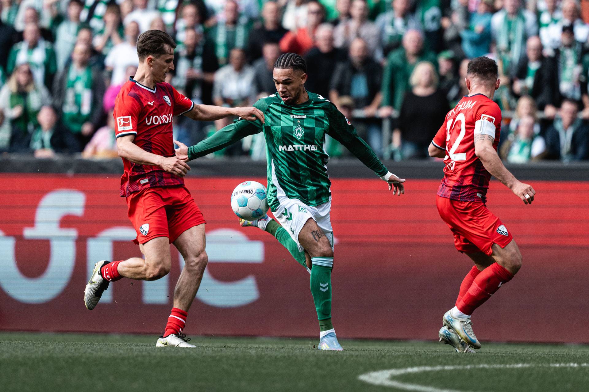 Fußballspiel, bei dem ein Spieler in Grün den Ball unter Kontrolle hat, während zwei Spieler in Rot ihn bekämpfen – vor einer jubelnden Zuschauermenge.