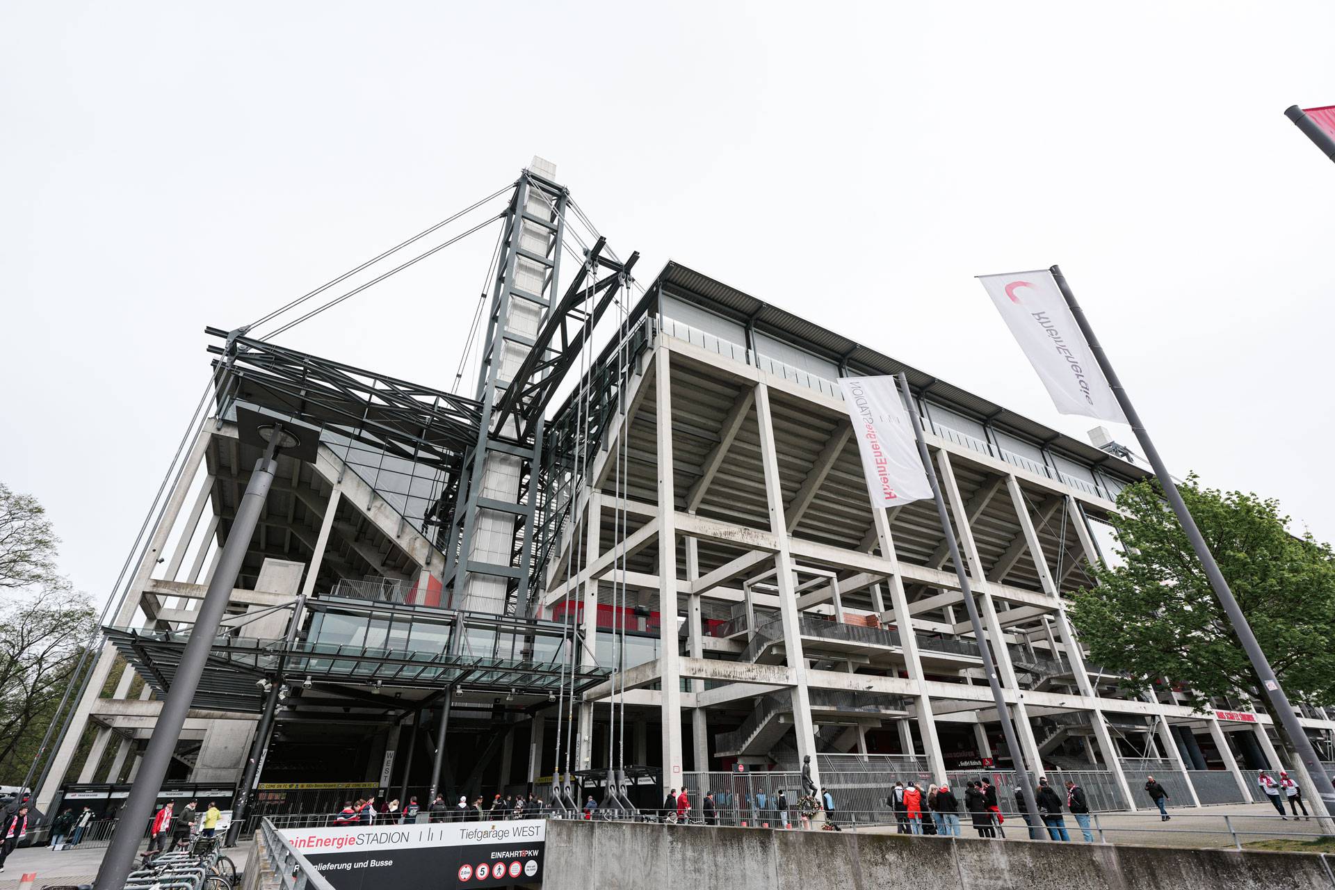 Außenansicht eines großen, modernen Stadions mit Metall- und Betonkonstruktionen, Menschen, die sich am Eingang versammelt haben, und wehende Fahnen vor dem Hintergrund eines bewölkten Himmels.