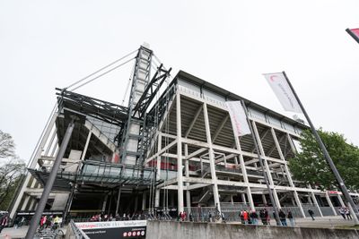 Außenansicht eines großen, modernen Stadions mit Metall- und Betonkonstruktionen, Menschen, die sich am Eingang versammelt haben, und wehende Fahnen vor dem Hintergrund eines bewölkten Himmels.
