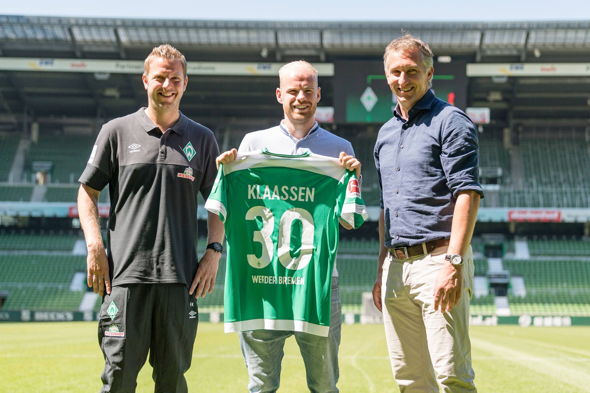 Drei Männer stehen in einem Stadion, wobei die Person in der Mitte ein grünes Fußballtrikot mit dem Namen „Klaassen“ und der Nummer 30 in der Hand hält.