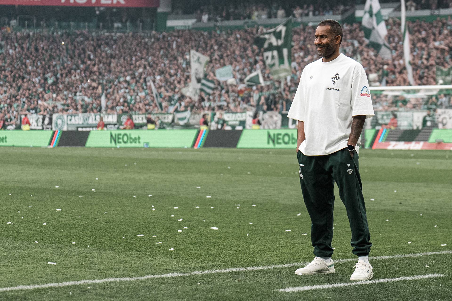 Ein Mann in weißem Hemd und dunkler Hose steht lächelnd auf dem Fußballplatz. Im Hintergrund füllen Fans mit grünen Transparenten das Stadion.