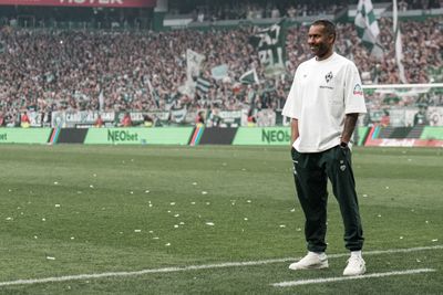 Ein Mann in weißem Hemd und dunkler Hose steht lächelnd auf dem Fußballplatz. Im Hintergrund füllen Fans mit grünen Transparenten das Stadion.
