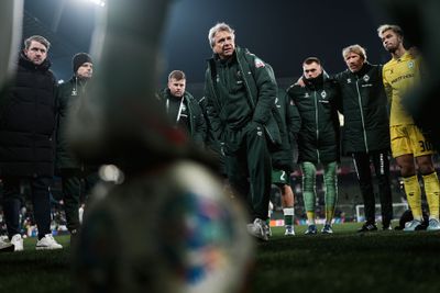 Trainer in grüner Jacke spricht zu Fußballmannschaft, die im Kreis auf dem Spielfeld steht, Spieler hören aufmerksam zu, Stadionbeleuchtung im Hintergrund.
