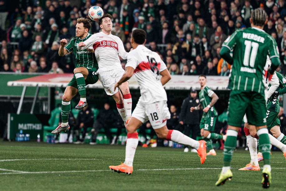 Fußballspieler in einem grün-weißen Trikot und weiß-roten Trikot kämpfen während eines Spiels um einen Kopfball, während im Hintergrund die Zuschauer zuschauen.