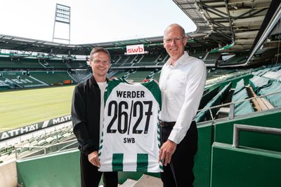 Zwei Männer stehen in einem Stadion und halten ein Werder-Trikot mit der Nummer 26/27 in den Händen.