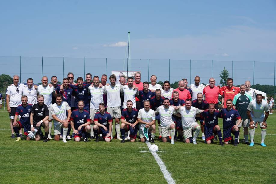 Zwei Fußballmannschaften posieren gemeinsam auf einem grünen Rasen unter strahlend blauem Himmel und tragen dunkelblaue und weiße Trikots.