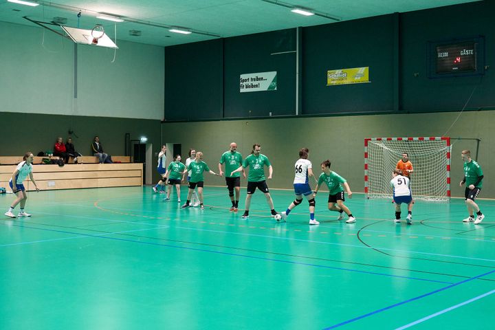 Handballspiel mit Spielern in grünen und weißen Trikots auf einem türkisfarbenen Spielfeld. Im Hintergrund sitzen Zuschauer auf Holzbänken.