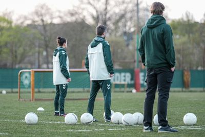 Drei Personen in Sportkleidung stehen auf einem Fußballplatz, auf dem mehrere weiße Fußbälle verstreut liegen.