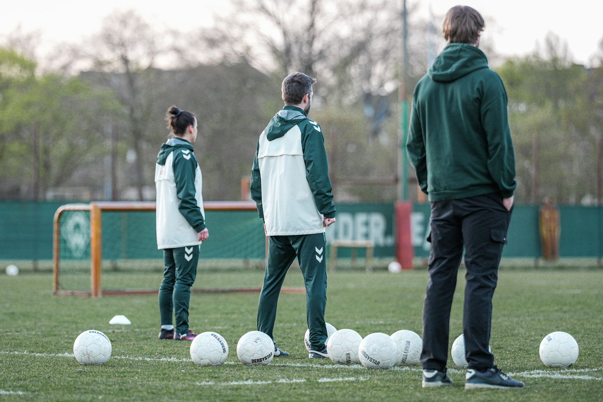 Drei Personen in Sportkleidung stehen auf einem Fußballplatz, auf dem mehrere weiße Fußbälle verstreut liegen.