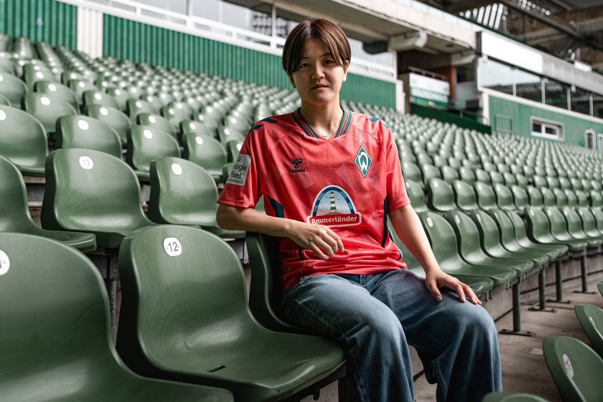 Eine Person in einem roten Werder-Bremen-Trikot sitzt zwischen leeren grünen Stadionplätzen und blickt in die Kamera.