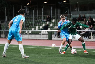 Fußballspieler in Aktion während eines Spiels; ein Spieler in Grün verteidigt den Ball gegen zwei Spieler in Blau auf einem Rasenfeld.