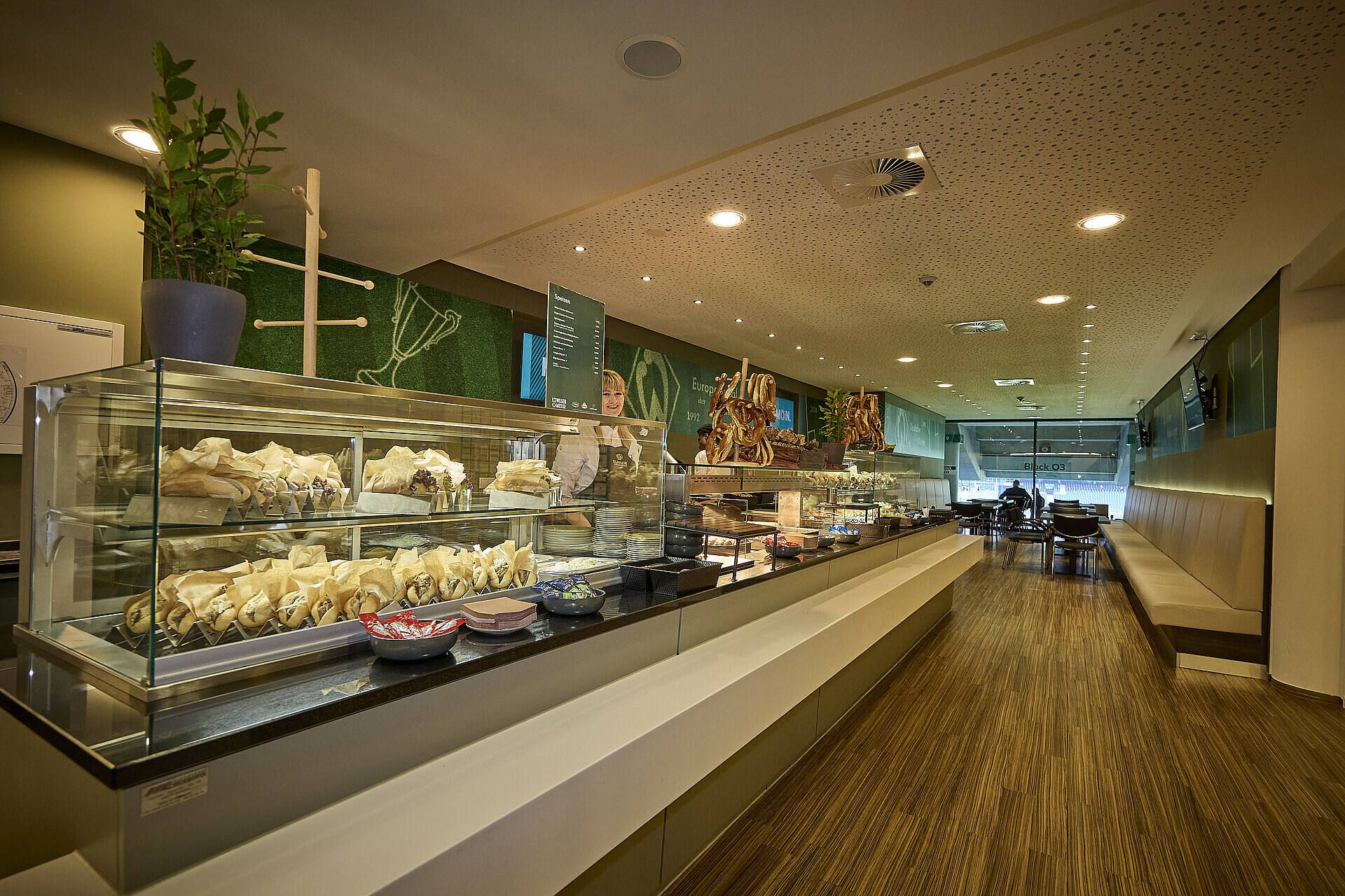 Modernes Café-Interieur mit einer langen Ausstellungsvitrine, in der Gebäck, Brot und Snacks präsentiert werden. Holzfußboden und Sitzbereich mit Tischen im Hintergrund.