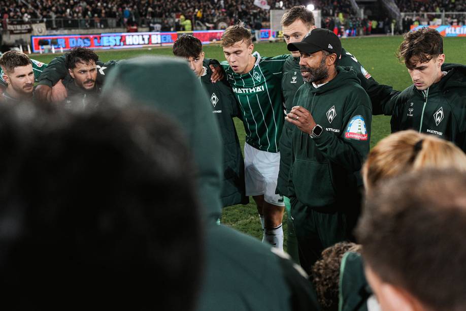 Ein Fußballtrainer hält eine motivierende Ansprache an die Spieler in grünen Trikots, die sich auf dem Spielfeld versammelt haben, während im Hintergrund eine Menschenmenge zu sehen ist.
