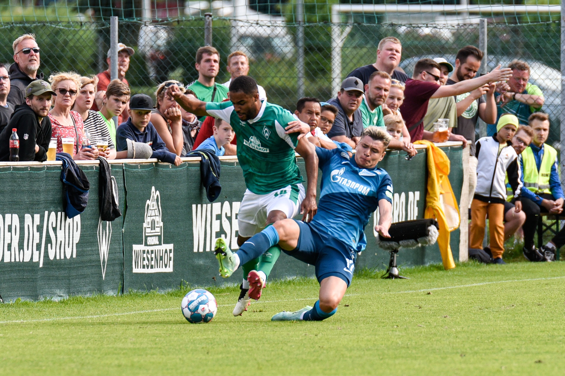 Ein Fußballspieler im grünen Trikot wird von einem anderen Spieler in Blau nahe der Seitenlinie zu Boden gebracht, während die Zuschauer auf den Tribünen das Geschehen aufmerksam verfolgen.