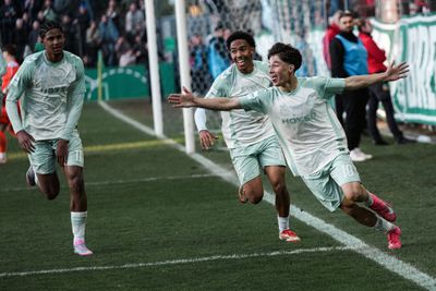 Drei begeisterte Fußballspieler in hellgrünen Trikots feiern auf einem Rasenfeld ein Tor, im Hintergrund jubeln Zuschauer.