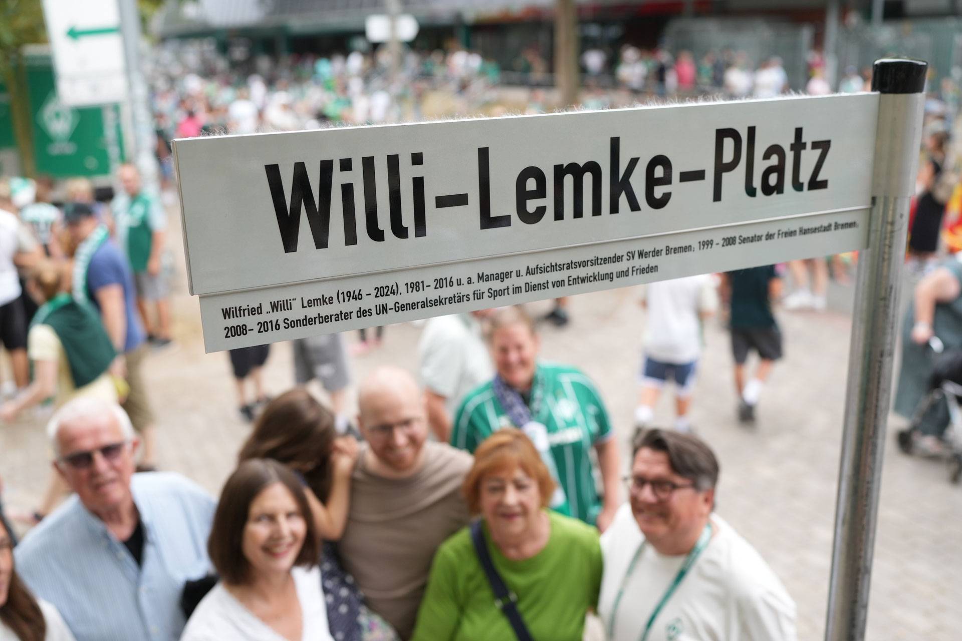 Ein Schild mit der Aufschrift „Willi-Lemke-Platz“, unter dem lächelnde Menschen stehen, in einem belebten Außenbereich.