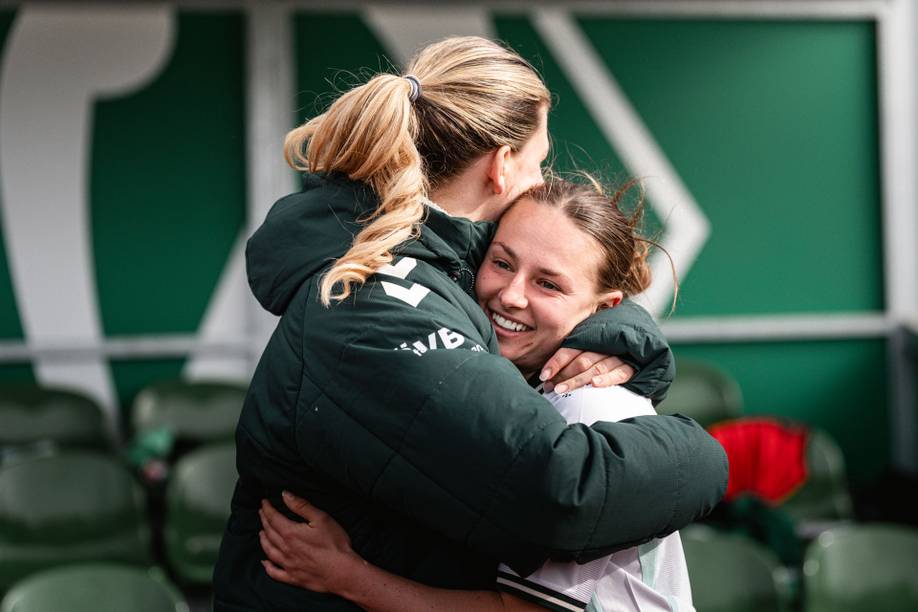Zwei Frauen, die sich freudig umarmen; eine von ihnen lächelt strahlend vor einer Sportkulisse mit grünen Sitzen und Hintergrund.