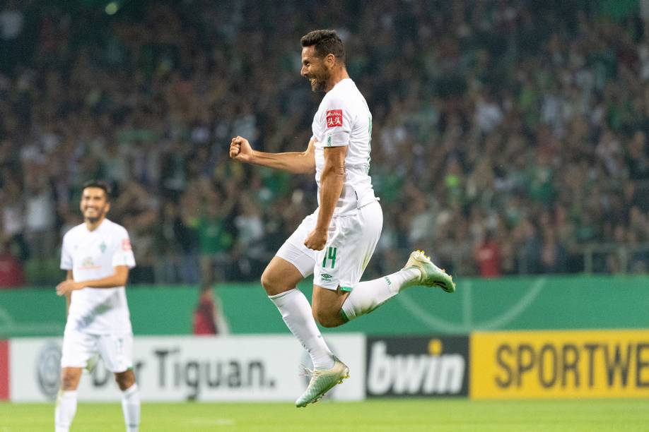 Ein Fußballspieler in weißem Trikot feiert ein Tor, springt mit geballter Faust in die Luft, im Hintergrund lächelt die Menge.