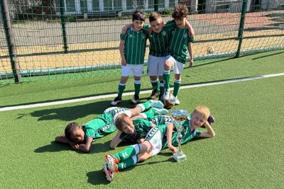 Vier junge Fußballspieler in grünen Trikots stehen lächelnd da, während andere mit einem Pokal auf dem Rasen liegen und triumphierend feiern.
