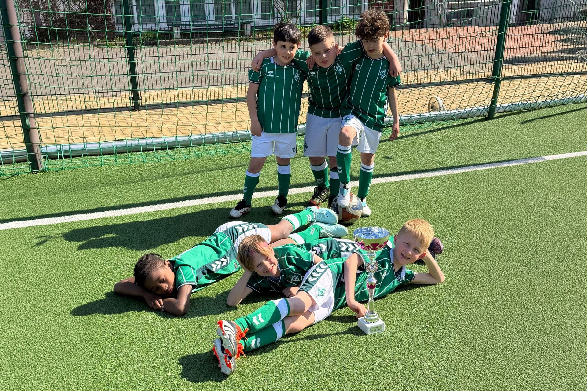 Vier junge Fußballspieler in grünen Trikots stehen lächelnd da, während andere mit einem Pokal auf dem Rasen liegen und triumphierend feiern.