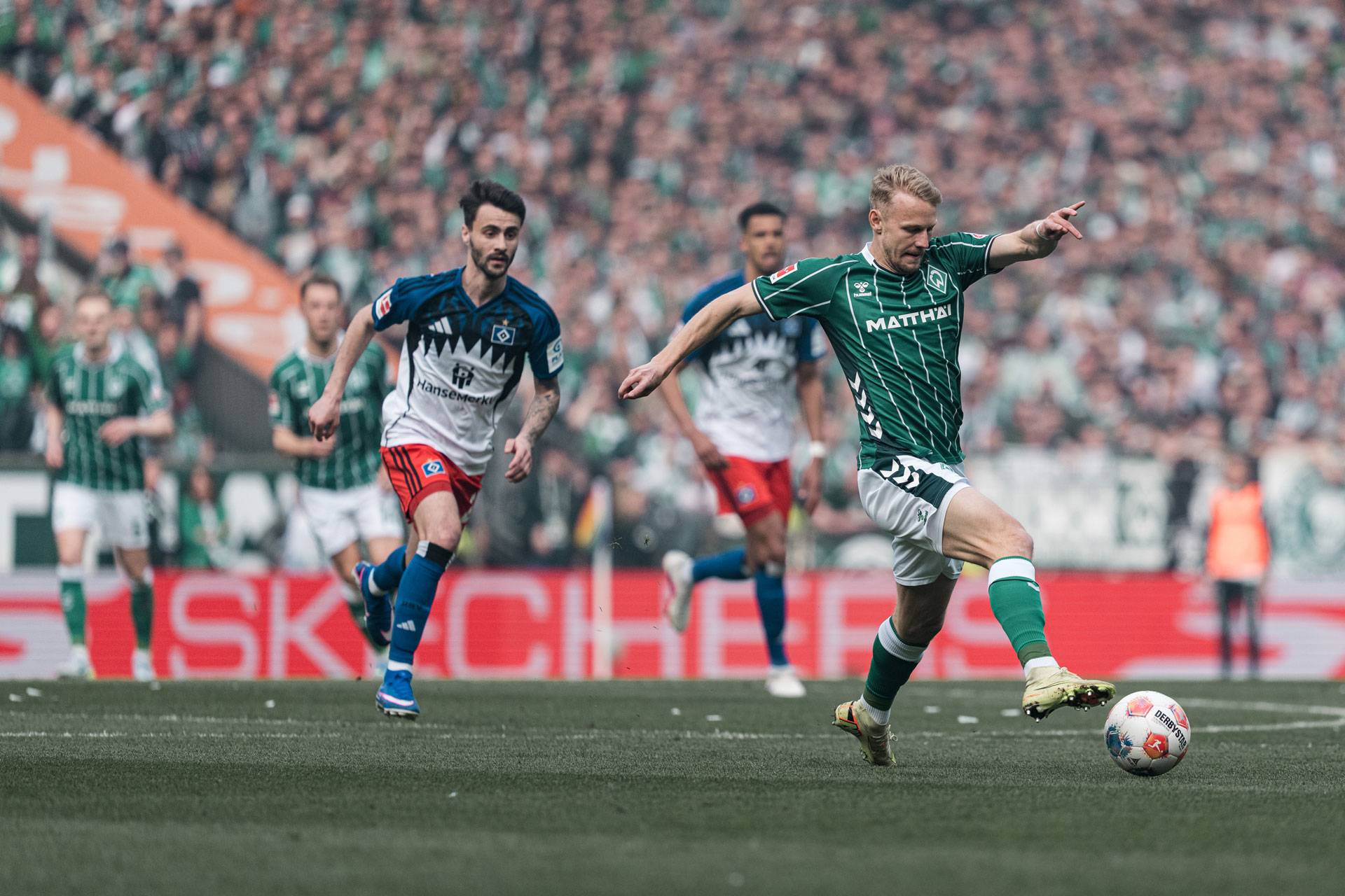 Ein Fußballspieler in einem grünen Trikot schießt während eines Spiels den Ball, im Hintergrund sind Spieler der gegnerischen Mannschaft und eine verschwommene Zuschauermenge zu sehen.
