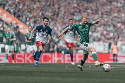 Ein Fußballspieler in einem grünen Trikot schießt während eines Spiels den Ball, im Hintergrund sind Spieler der gegnerischen Mannschaft und eine verschwommene Zuschauermenge zu sehen.