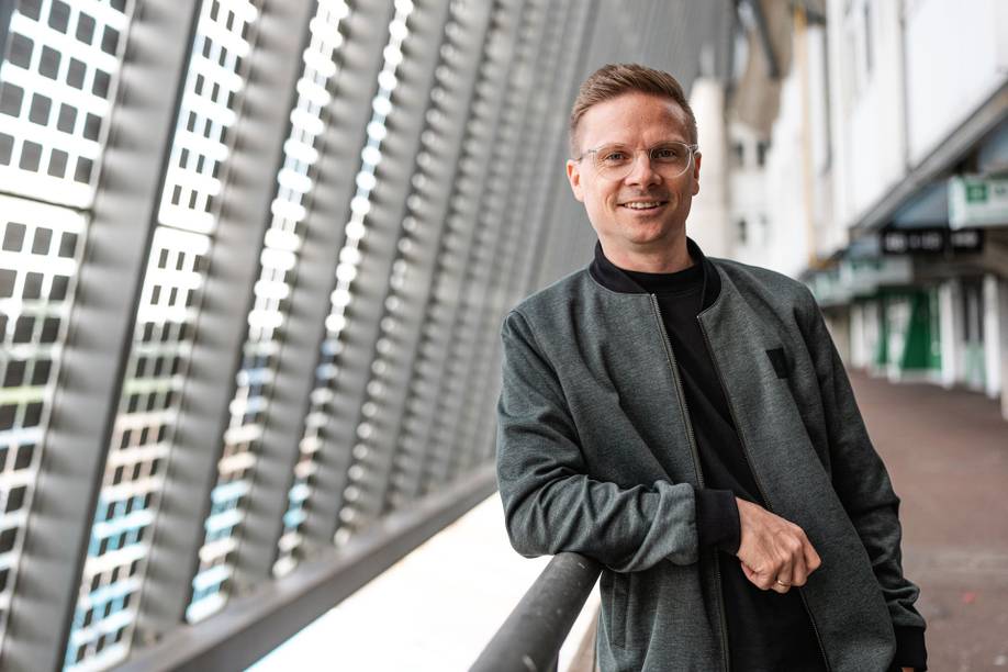 Ein Mann in einer dunklen Jacke steht lächelnd neben einem Geländer, im Hintergrund sind die gemusterten Fenster eines modernen Gebäudes zu sehen.