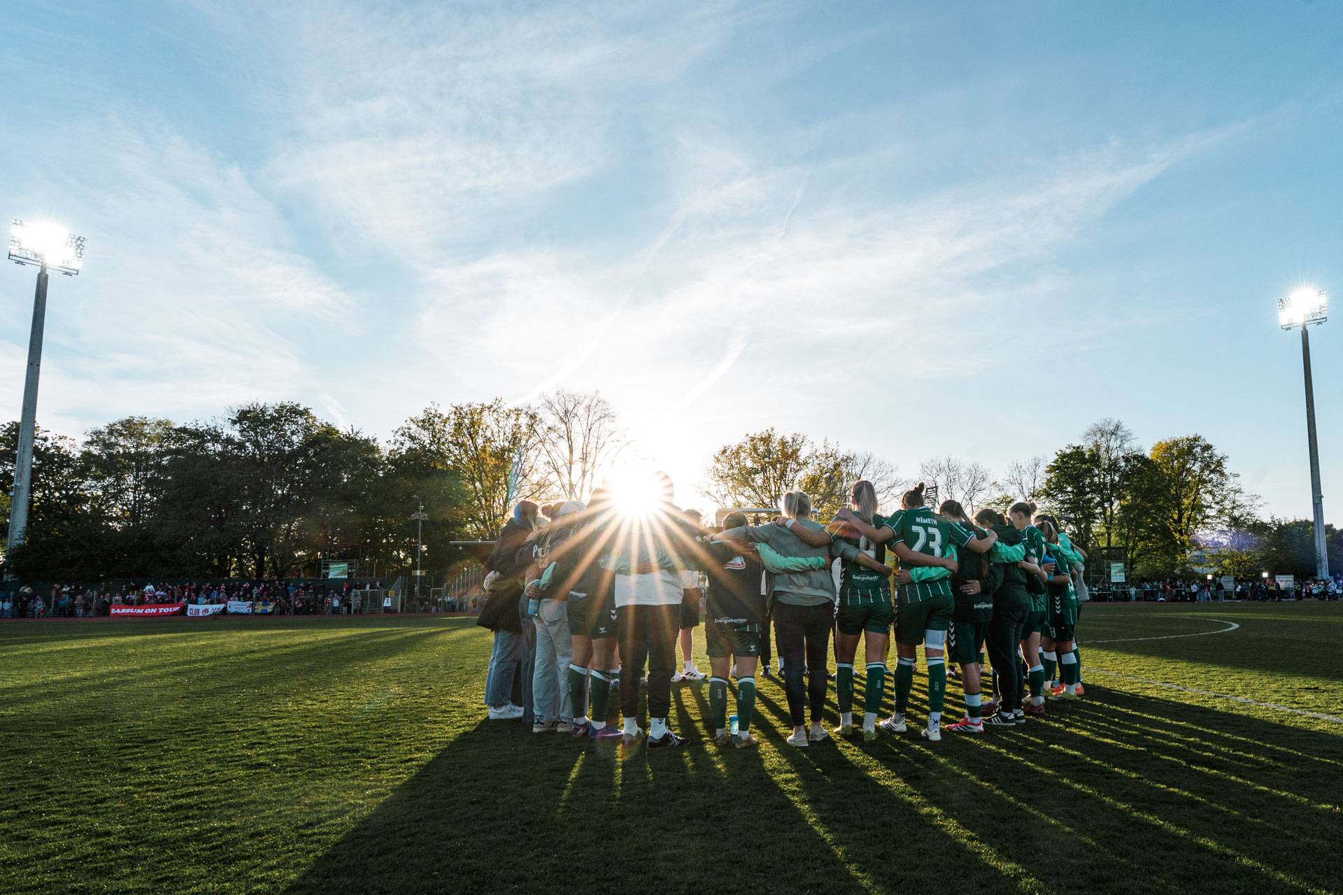 Eine Fußballmannschaft steht im Kreis auf einem Spielfeld, über das Sonnenlicht hereinströmt, im Hintergrund sind Bäume und Stadionbeleuchtung zu sehen.