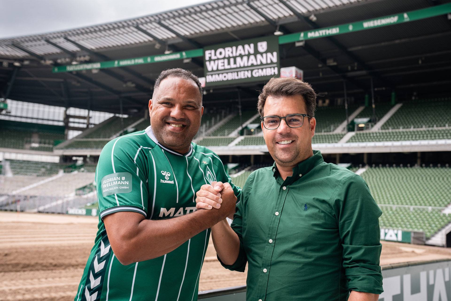 Zwei Männer, die in einem Stadion lächeln und sich die Hand geben; beide tragen grüne Trikots. Im Hintergrund sind Stadionränge und Werbebanner zu sehen.