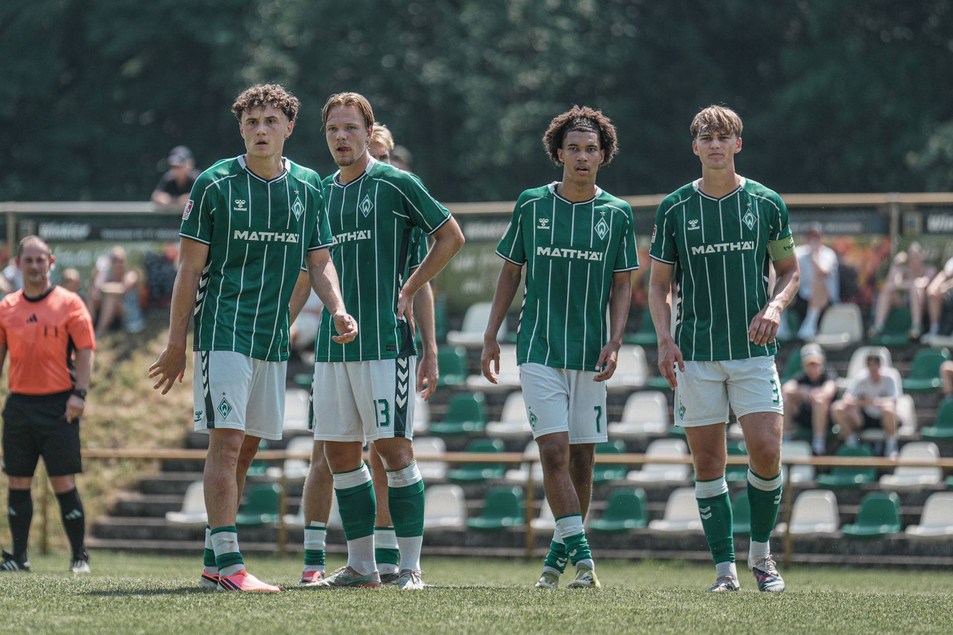Vier Fußballspieler in grün-weißen Trikots stehen auf dem Spielfeld, im Hintergrund sind ein Schiedsrichter und Zuschauer zu sehen.